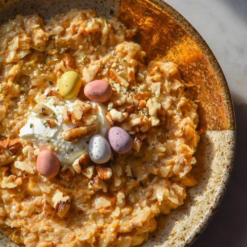 An aerial image of a beige speckled ceramic bowl filled with gluten free carrot cake porridge. The porridge is topped with chopped walnuts, speckled eggs and yoghurt and sits on a white marble table in contrasting sunlight.