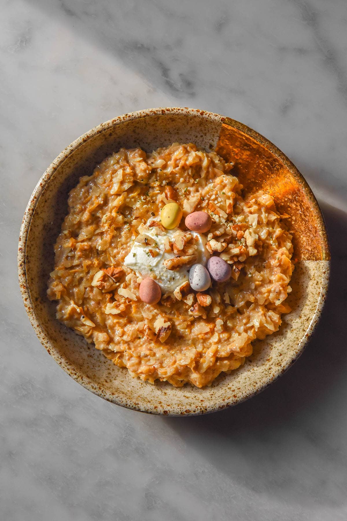 An aerial image of a beige speckled ceramic bowl filled with gluten free carrot cake porridge. The porridge is topped with chopped walnuts, speckled eggs and yoghurt and sits on a white marble table in contrasting sunlight.