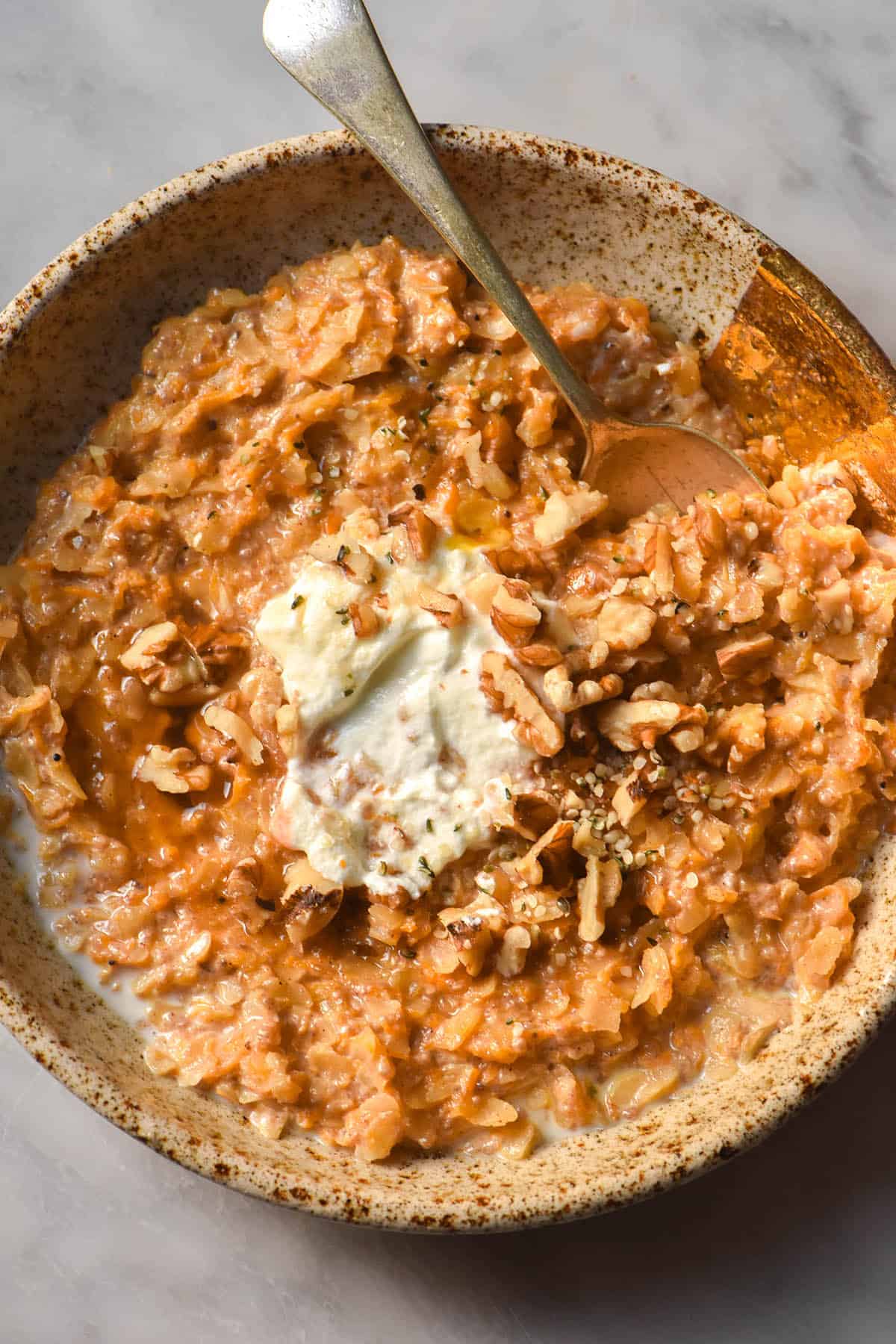 An aerial close up image of gluten free carrot cake porridge topped with Greek yoghurt, walnuts and hemp seeds on a white marble table.