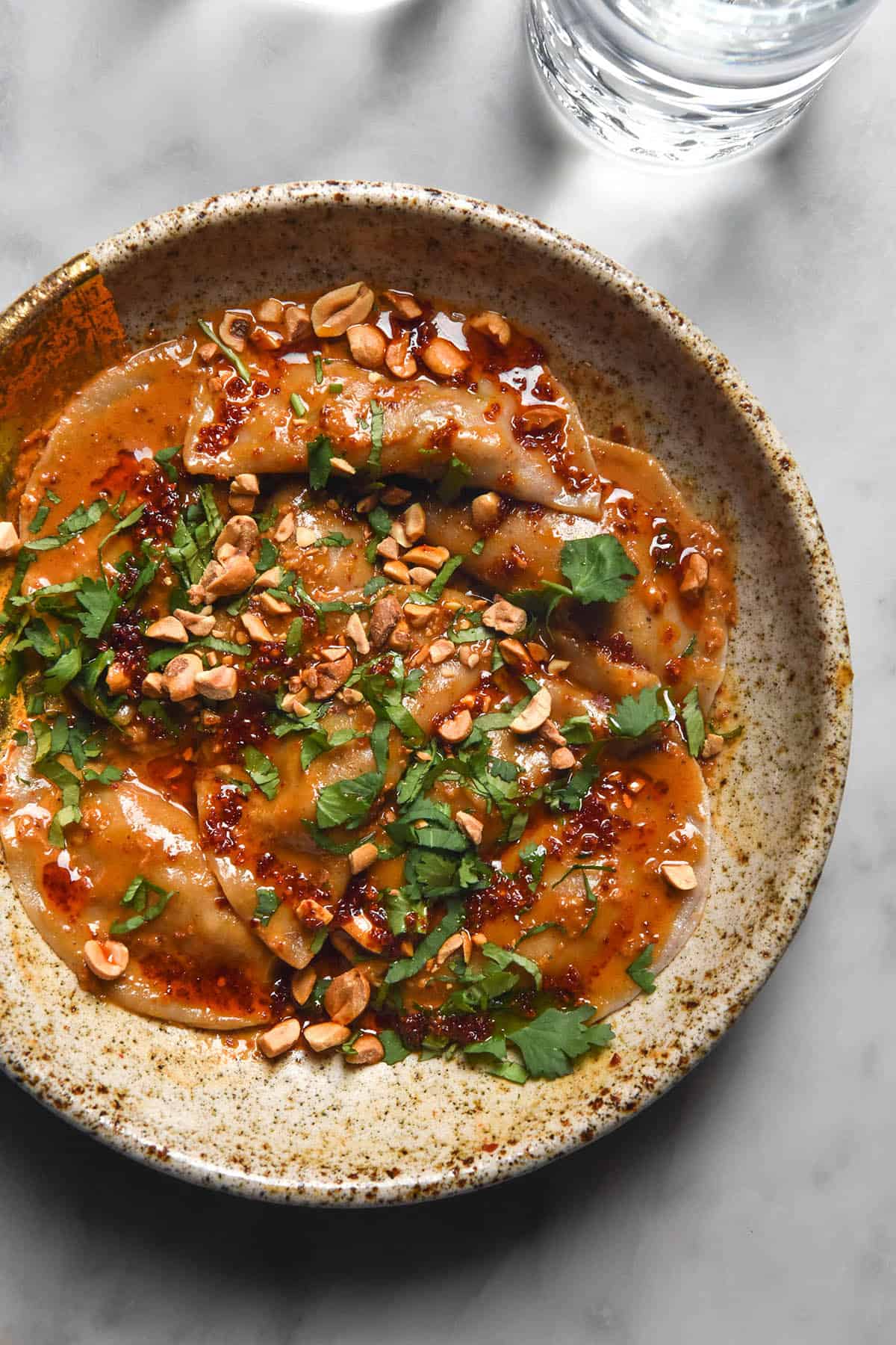 An aerial image of a beige speckled ceramic bowl filled with gluten free dumplings in a peanut chilli oil sauce and topped with coriander and chopped peanuts. The bowl sits atop a white marble table and two sunlit glasses of water sit to the top of the image.