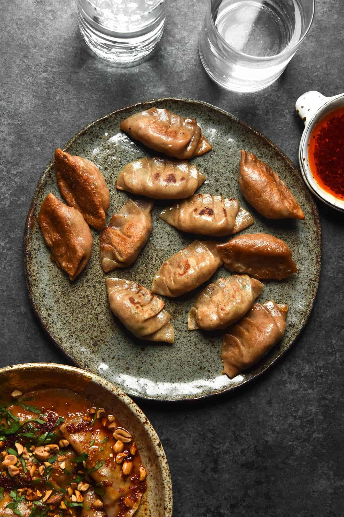 An aerial image of pleated and fried gluten free dumplings on a dark blue speckled ceramic plate atop a dark blue backdrop. The central plate is surrounded by water glasses, some chilli oil and a second bowl of dumplings in a peanut sauce. 