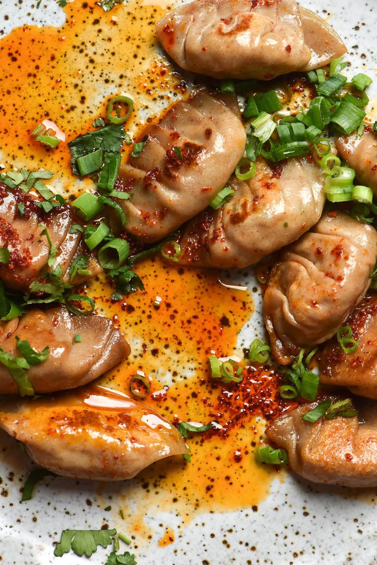 A close up aerial image of a white speckled ceramic plate topped with pleated dumplings smothered in chilli oil and topped with spring onion greens, coriander and chopped peanuts.