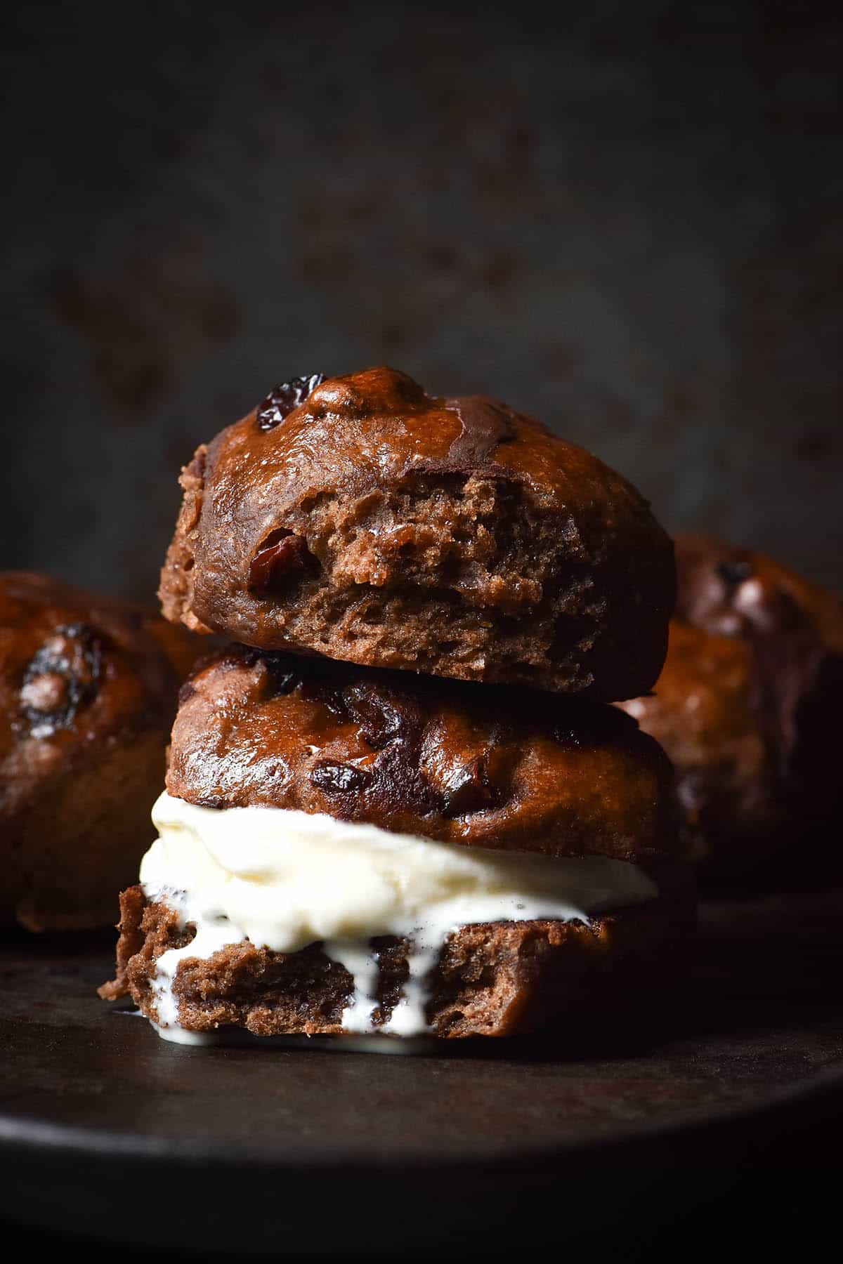 A moody side on image of a toasted gluten free hot cross bun sandwiched with vanilla ice cream. A second hot cross bun sits on top of it and two buns sit either side of the stack in the background.
