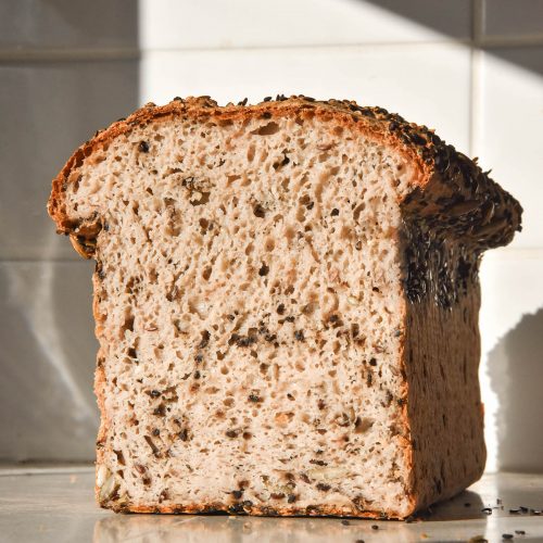 A brightly lit image of a loaf of gluten free seeded bread on a white bench top against a white stone wall. The loaf is filled with seeds and has been sliced to reveal the soft and fluffy inner crumb.