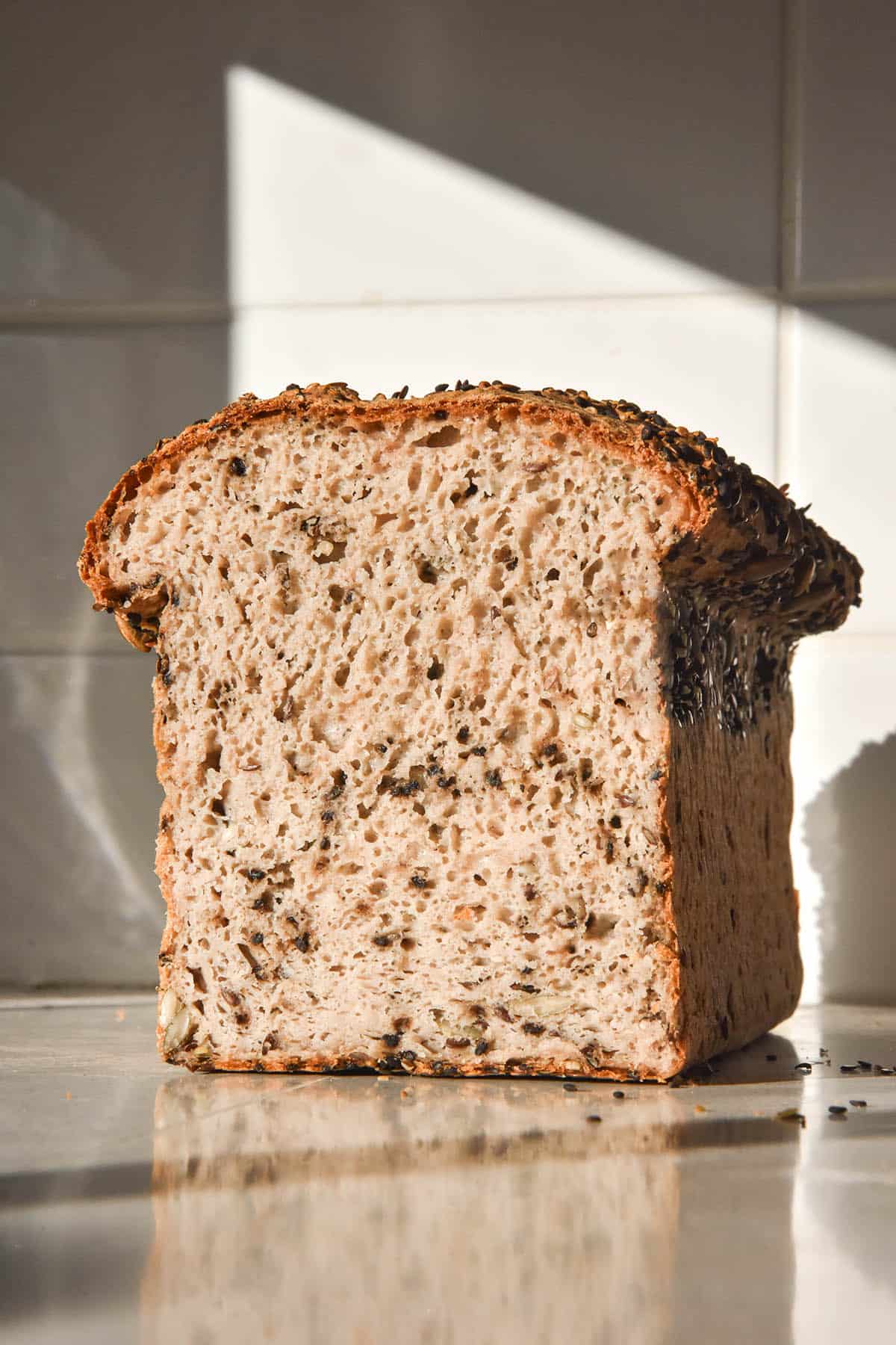 A brightly lit image of a loaf of gluten free seeded bread on a white bench top against a white stone wall. The loaf is filled with seeds and has been sliced to reveal the soft and fluffy inner crumb.