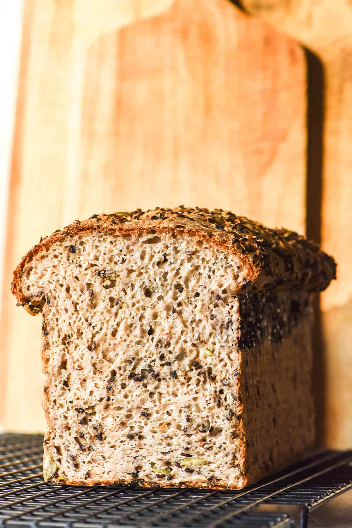 An image of a sliced loaf of gluten free seeded bread on a cooling rack against pale wood chopping boards in the background.