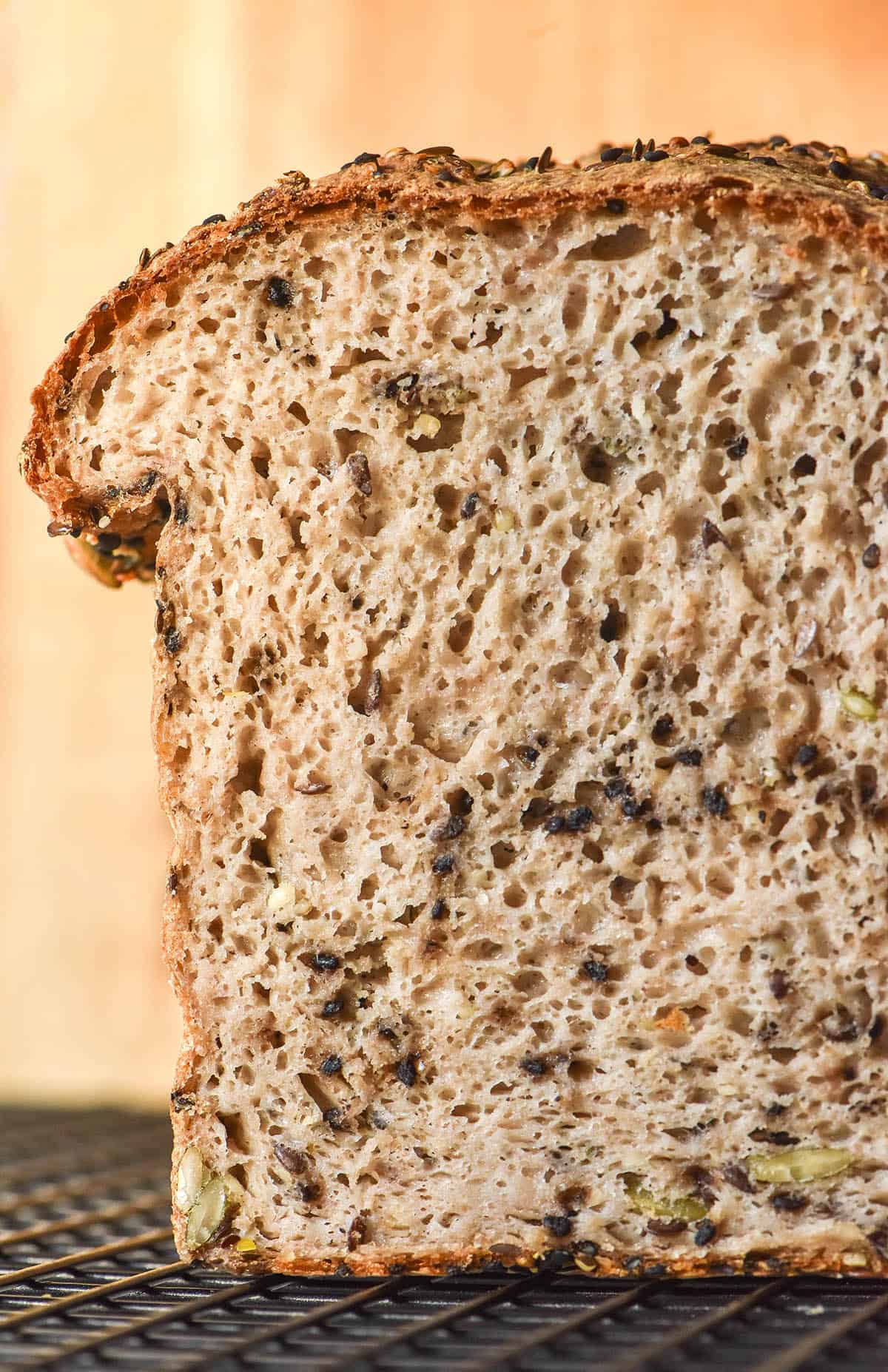 A side on close up image of a sliced loaf of gluten free seeded bread on a cooling rack set against a pale wood backdrop.
