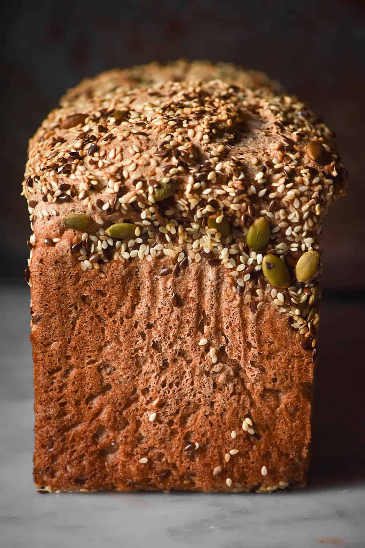 A moody side on image of a loaf of gluten free seeded bread on a white marble table against a dark backdrop