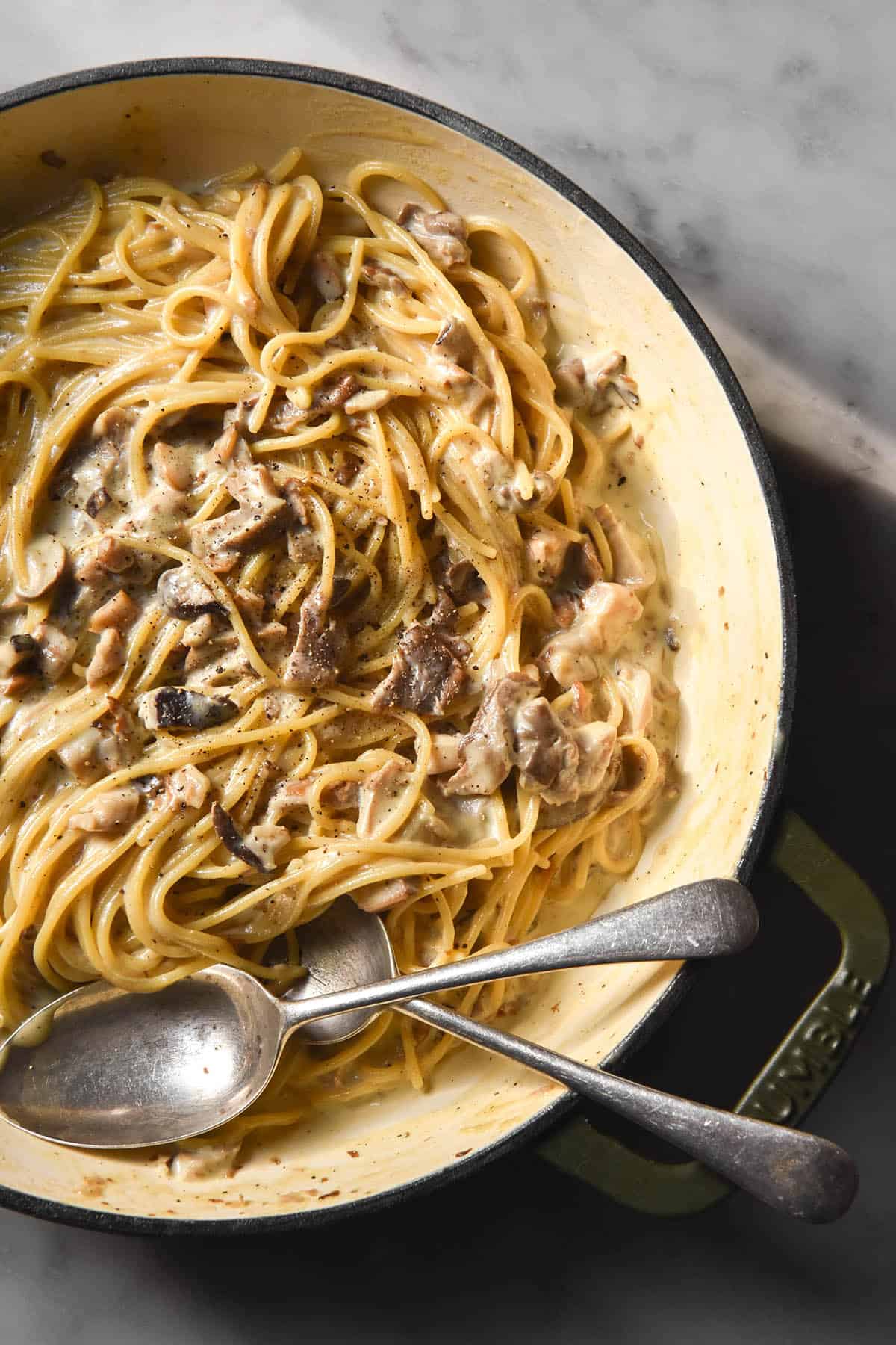An aerial image of a skillet filled with low FODMAP creamy mushroom pasta. The skillet sits in sunlight on a white marble table and two spoons stick out to the bottom left of the image.