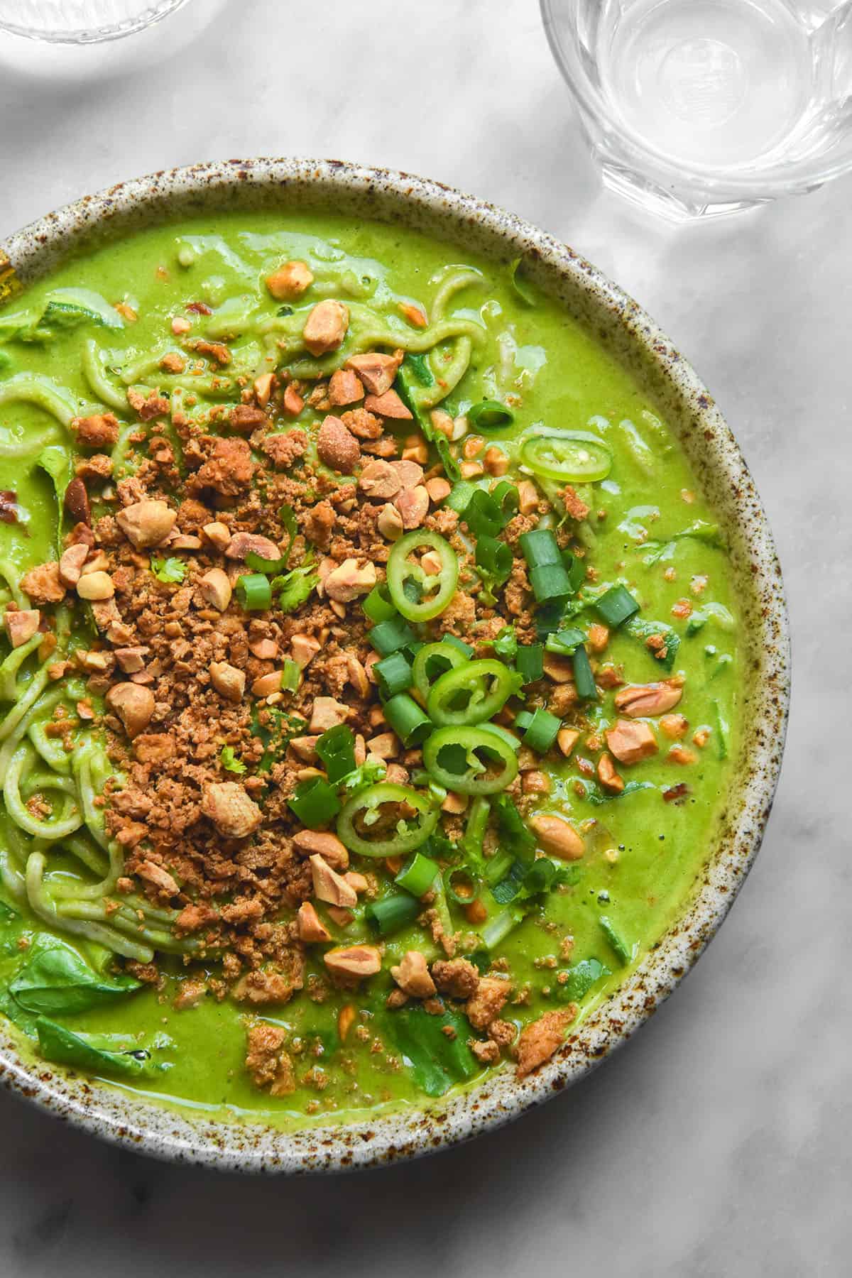 An aerial image of a bowl of low FODMAP green soup topped with tofu crumbles, peanuts, chopped green chilli and spring onion greens. The bowl sits atop a white marble table and two water glasses sit to the top of the image.