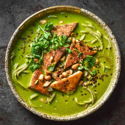 An aerial image of a bowl of low FODMAP green soup on a dark steel backdrop. The green soup is topped with cooked tofu triangles, peanuts, seeds and extra coriander. Noodles pot out from underneath the bright green broth.