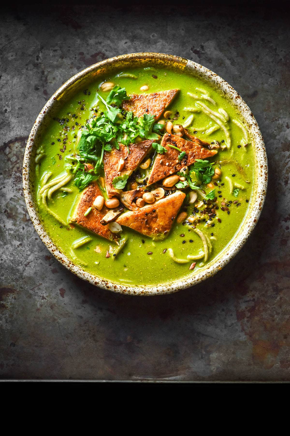 An aerial image of a bowl of low FODMAP green soup on a dark steel backdrop. The green soup is topped with cooked tofu triangles, peanuts, seeds and extra coriander. Noodles pot out from underneath the bright green broth.