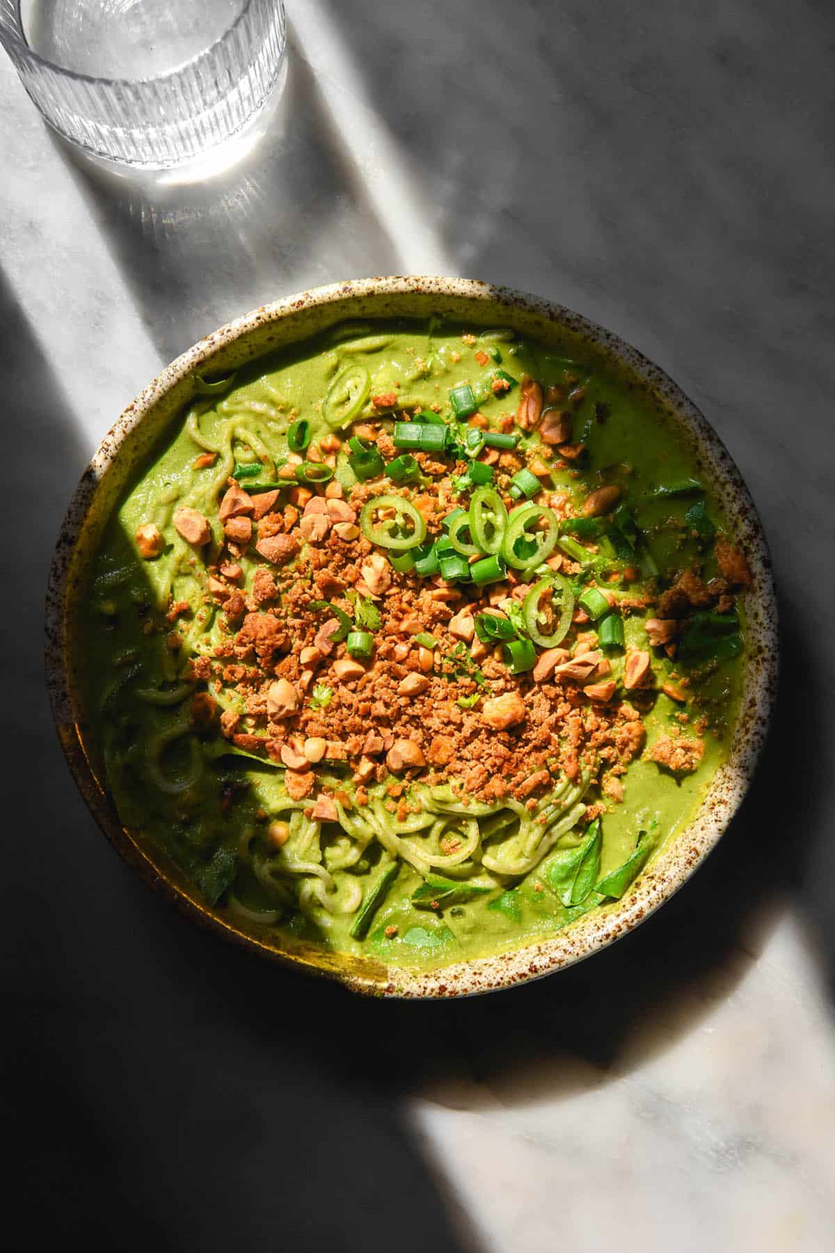 An aerial image of a bowl of low FODMAP green soup topped with tofu crumbles, peanuts, chopped green chilli and spring onion greens. The bowl sits atop a white marble table in deeply contrasting light and a water glass sit to the top of the image.