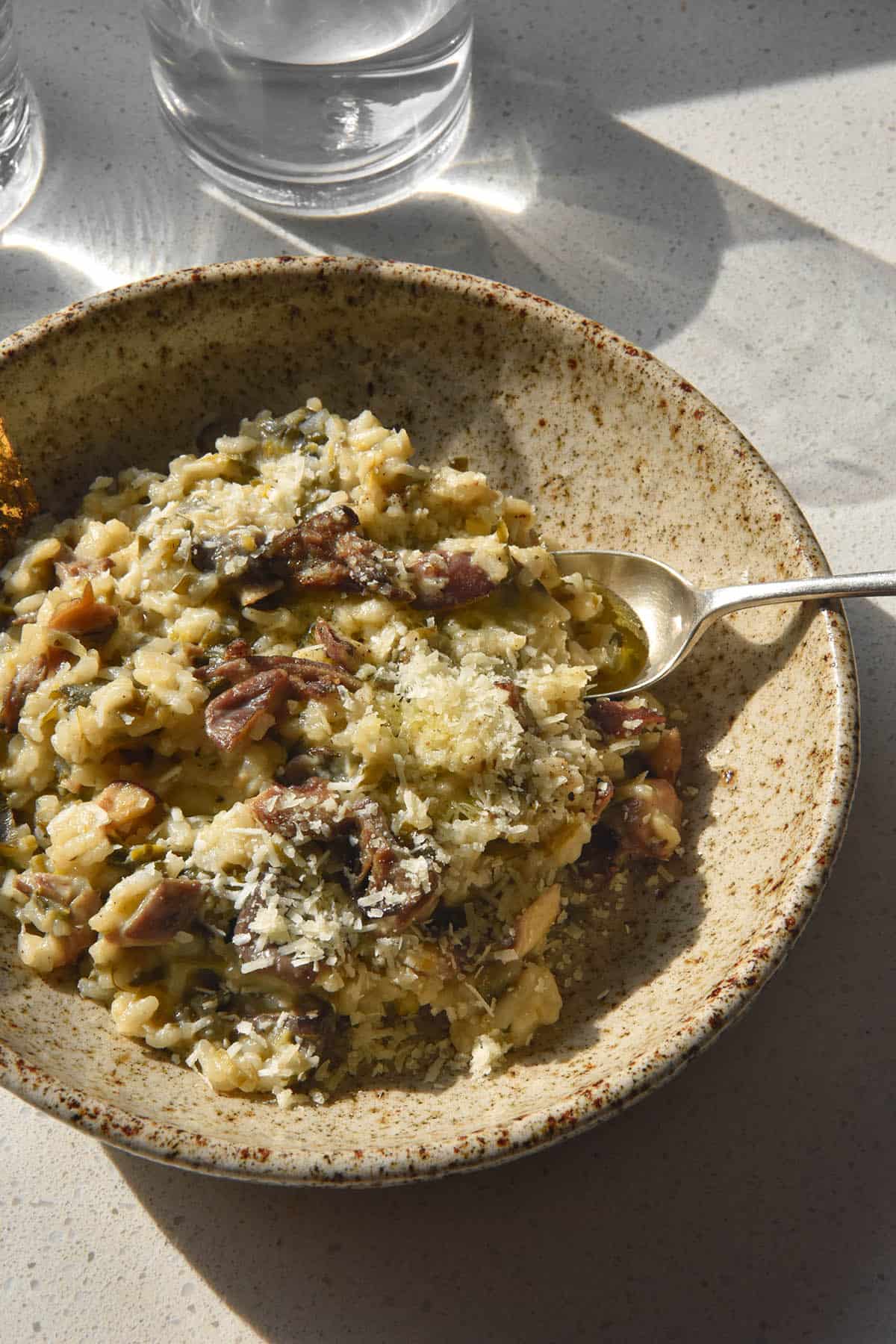 A close up side on view of a beige speckled ceramic bowl filled with low FODMAP mushroom risotto. The risotto is topped with grated parmesan and a spoon sits in the bowl extending to the right side of the image.