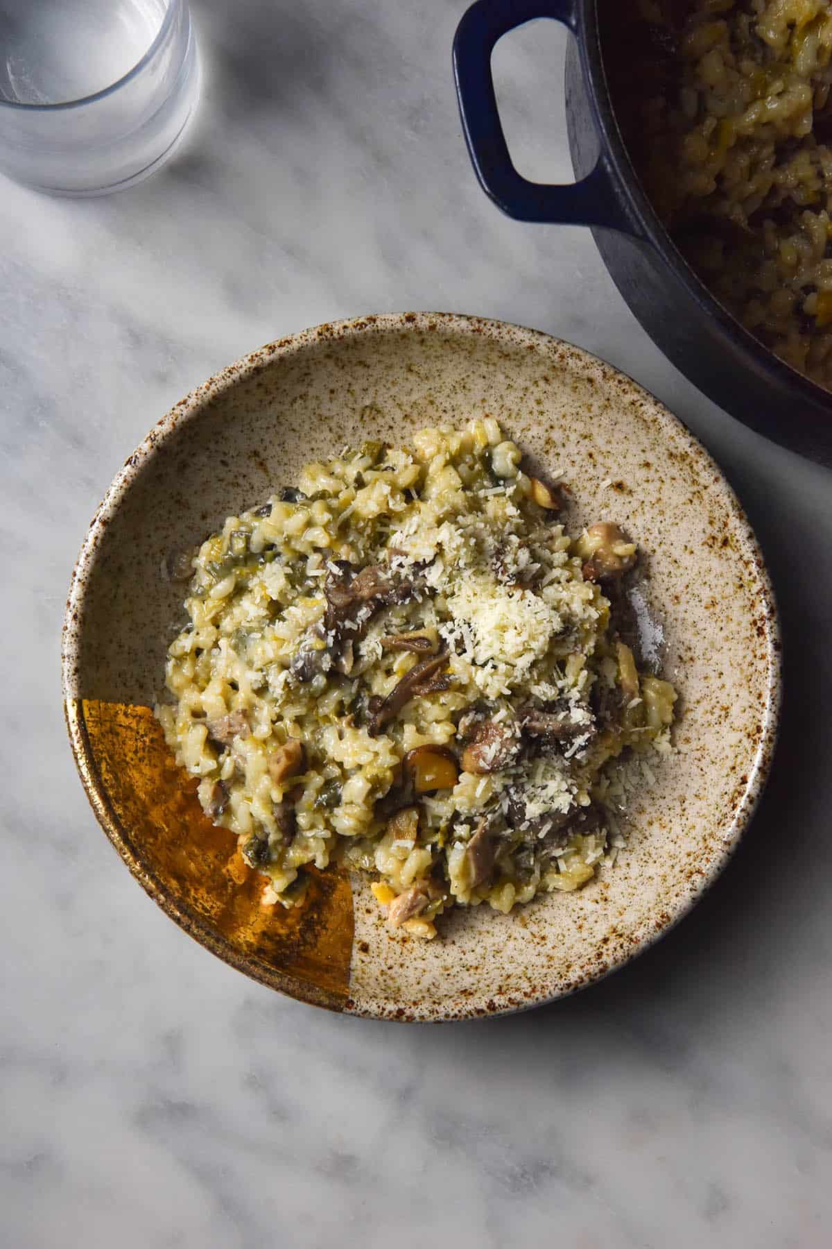 An aerial image of a bowl of low FODMAP mushroom risotto on a white marble table. A dutch oven filled with the remaining risotto sits to the right of the bowl, and a glass of water sits to the top left of the image.