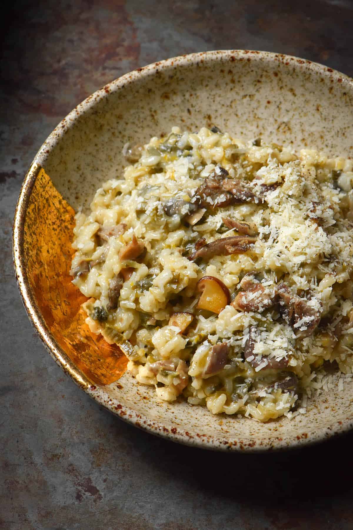 A close up side on view of a beige speckled ceramic bowl filled with low FODMAP mushroom risotto. The risotto is topped with grated parmesan and sits on a dark grey backdrop.