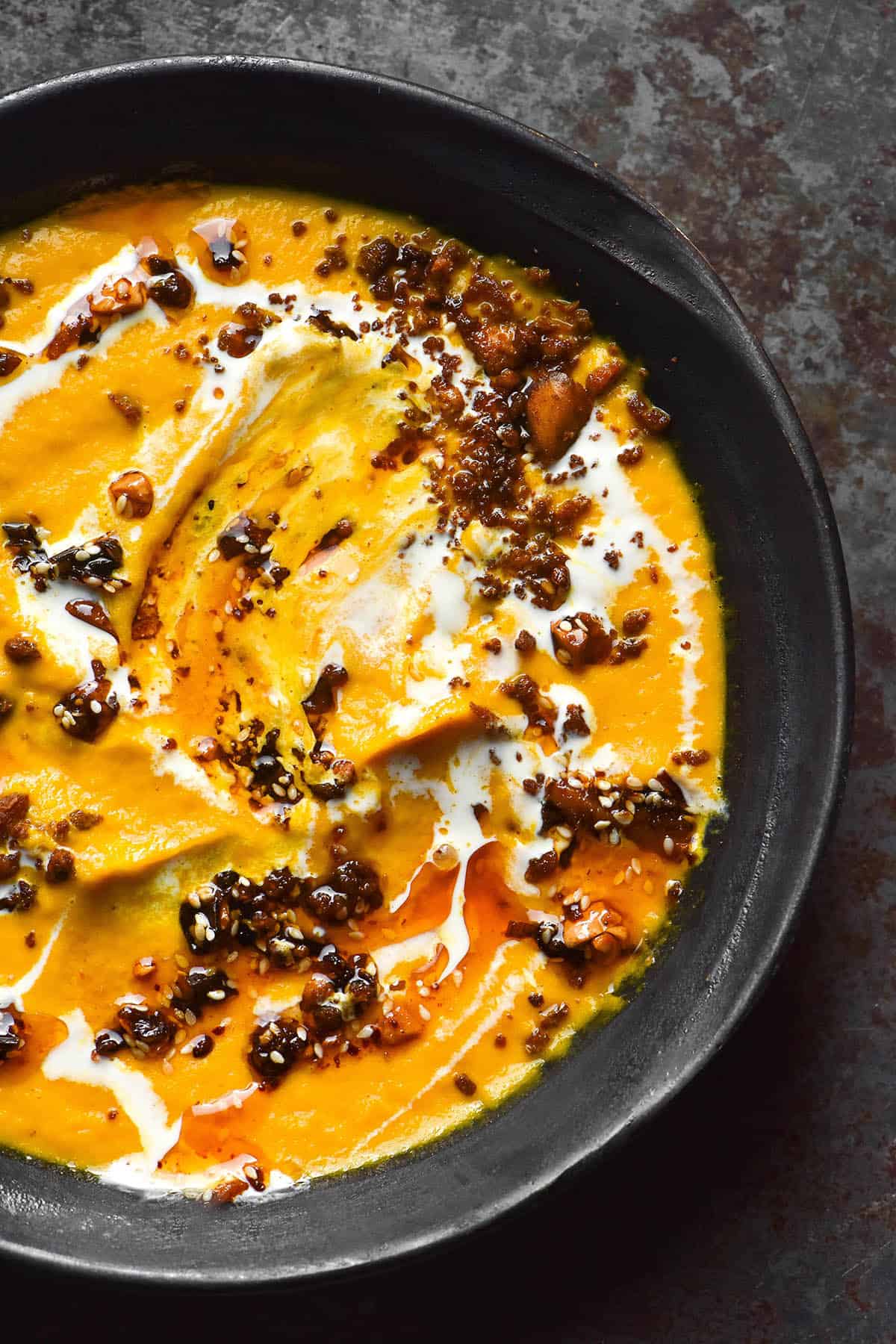 An aerial image of a dark grey ceramic bowl of carrot soup topped with tofu crumbles, cream and chilli oil on a dark metal backdrop.