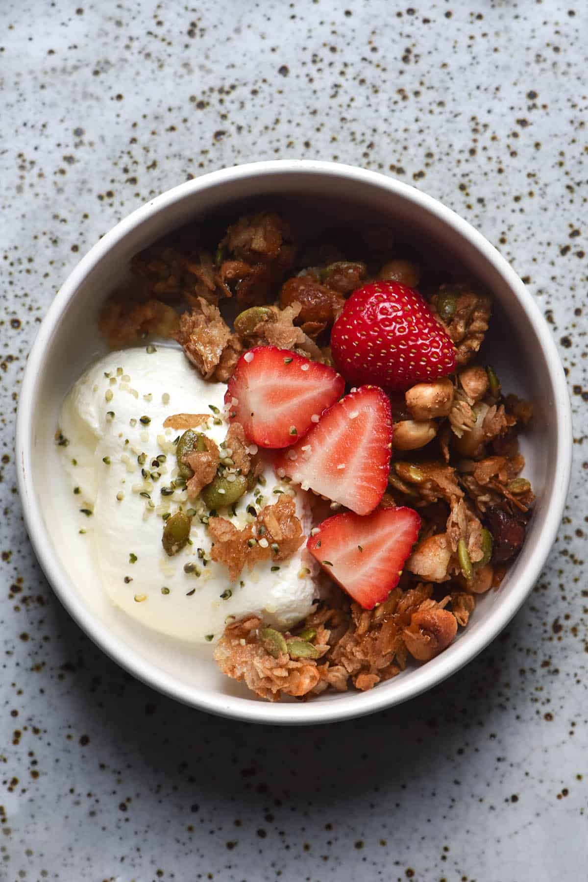 An aerial image of a white bowl filled with yoghurt, sliced strawberries and gluten free granola. The bowl sits atop a white speckled ceramic plate.