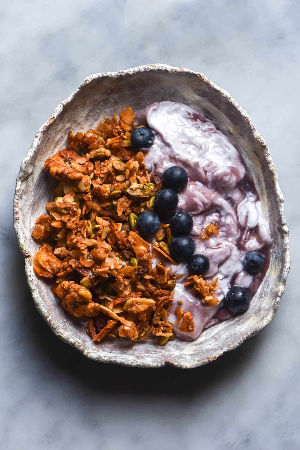 An aerial image of a white misshapen ceramic bowl on a white marble table filled with blueberry yoghurt and granola and topped with fresh blueberries.