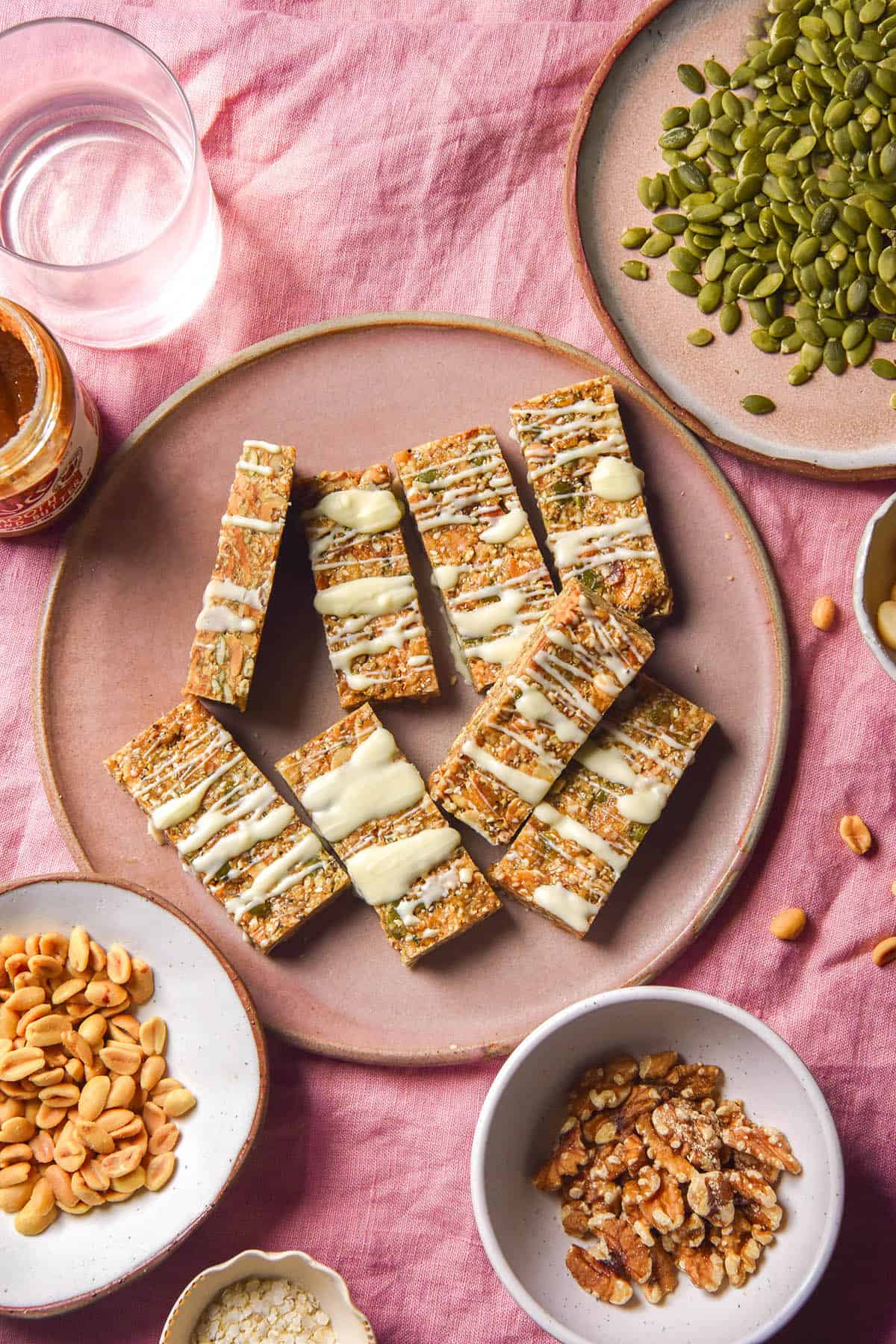 An aerial image of a pale pink ceramic plate of gluten free granola bars drizzled with white chocolate. The plate sits atop a pale pink linen tablecloth and is surrounded by small bowls of nuts and seeds and a glass of water