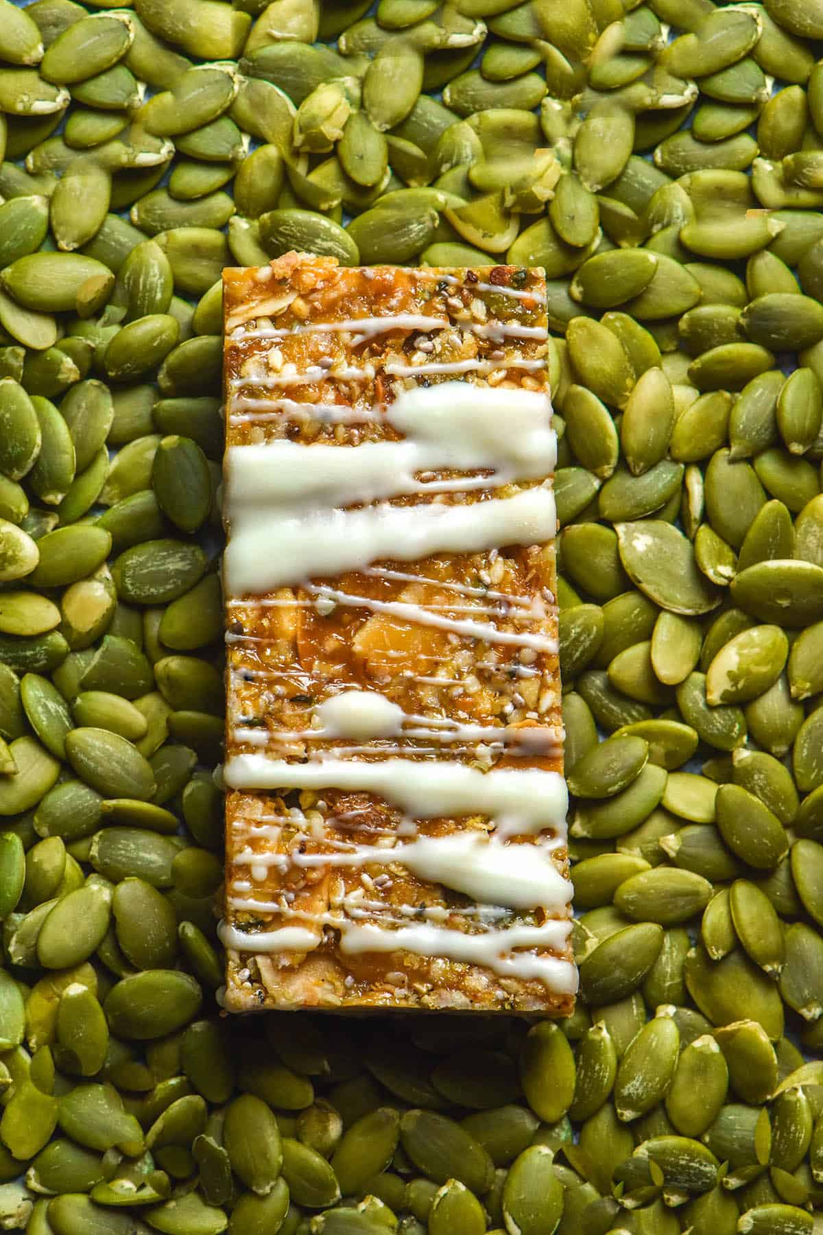 An aerial image of a gluten free granola bar drizzled with white chocolate sitting atop a plate full of pumpkin seeds