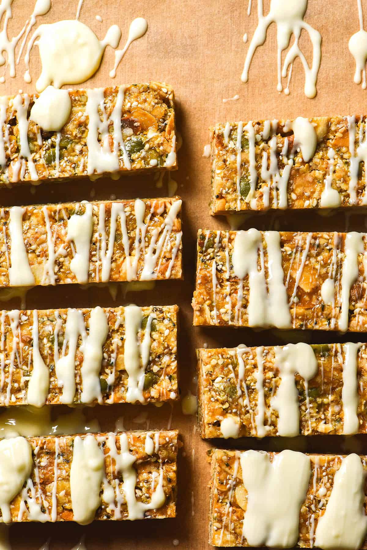 An aerial image of gluten free granola bars on a sheet of brown baking paper being drizzled with white chocolate