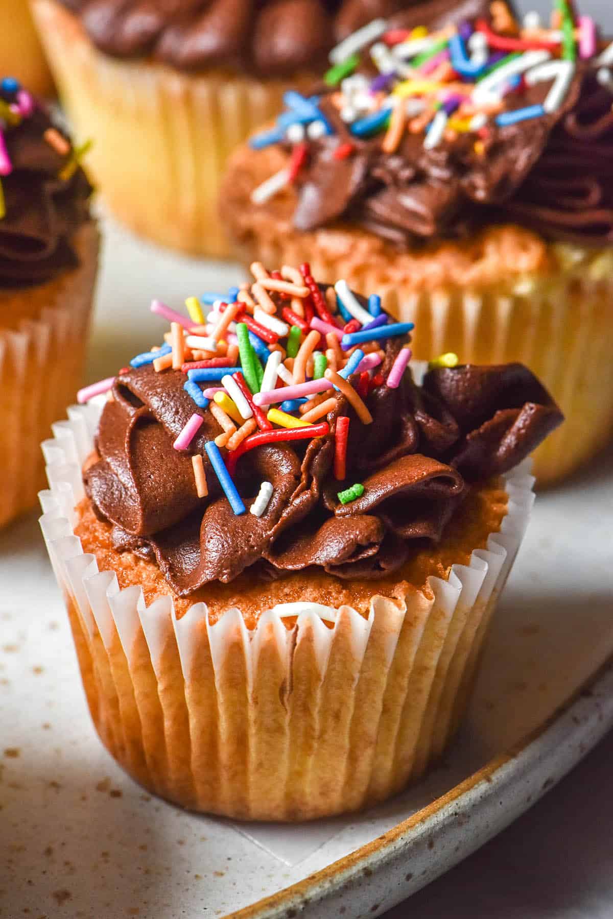 A side on view of gluten free vanilla cupcakes topped with chocolate buttercream and rainbow sprinkles
