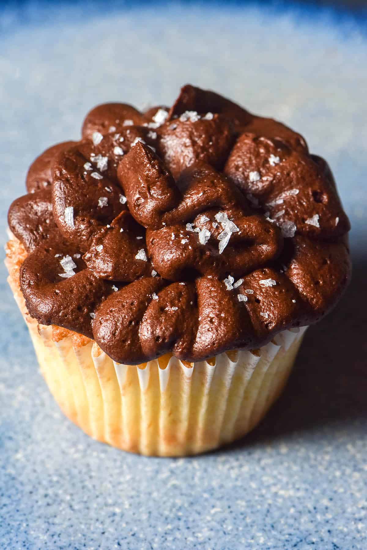 An aerial close up image of a gluten free vanilla cupcake topped with swirly chocolate buttercream and sea salt flakes atop a bright blue ceramic plate