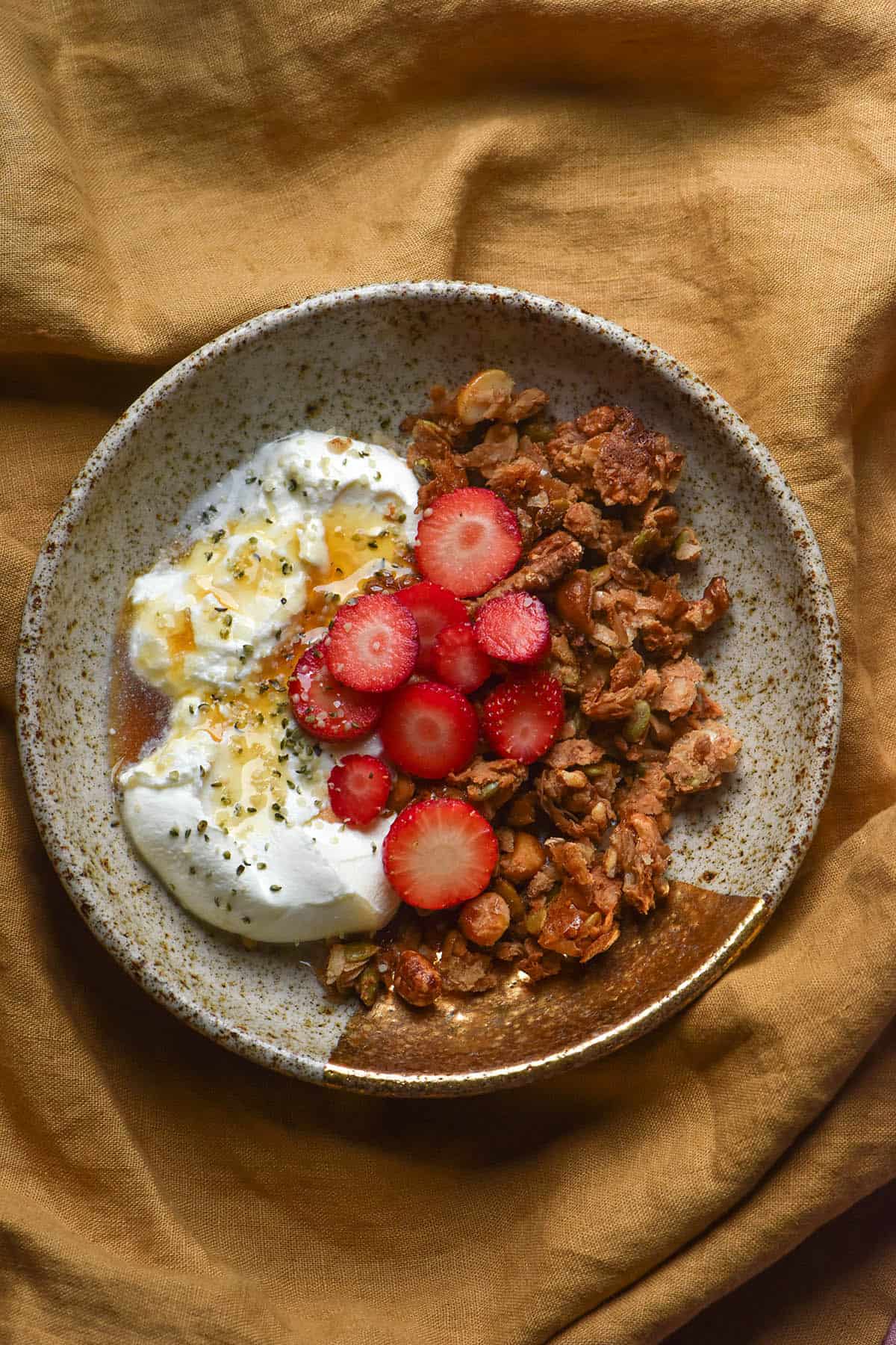 An aerial image of a beige ceramic bowl filled with low FODMAP granola, yoghurt and sliced strawberries atop a mustard linen tablecloth