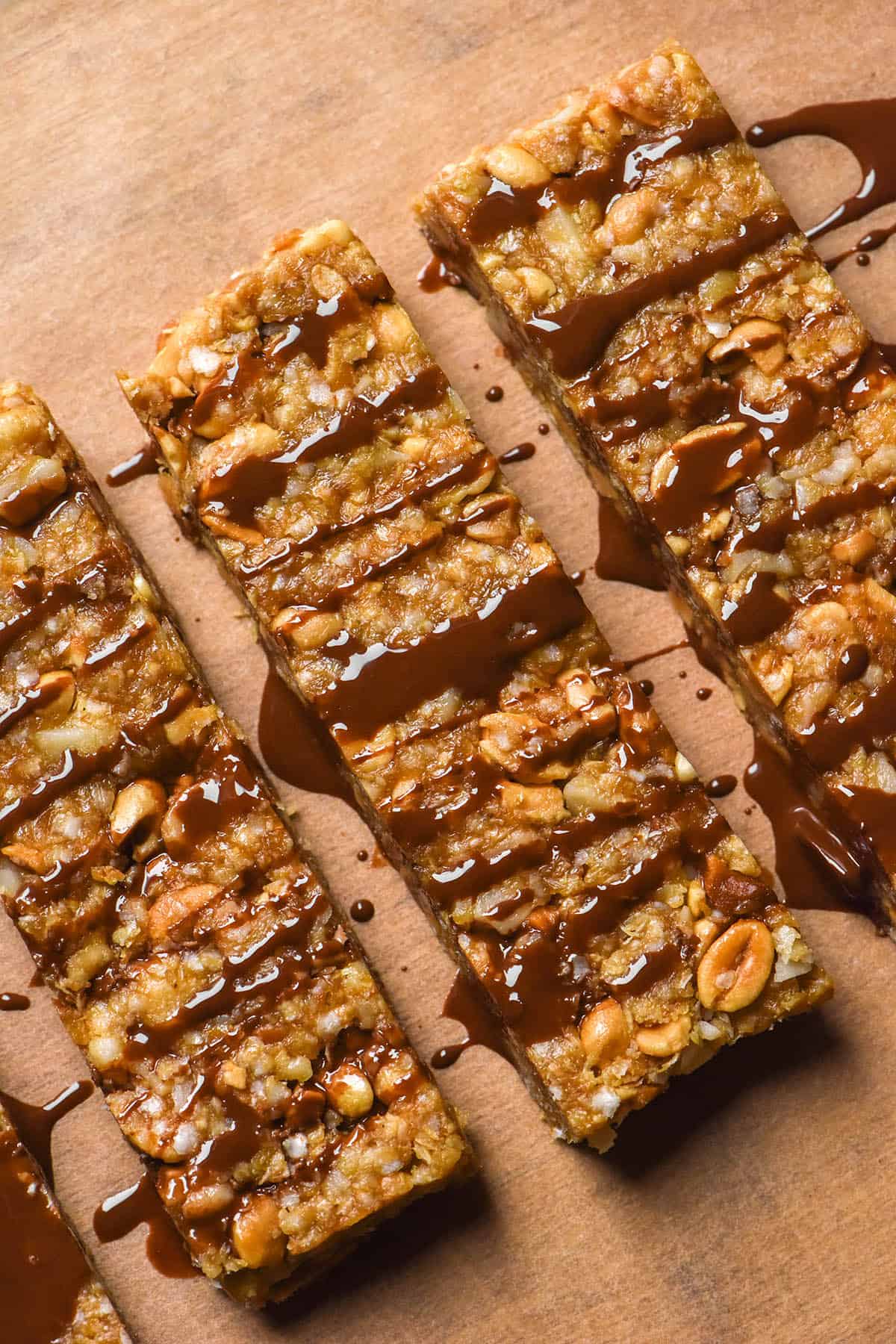 An aerial image of Low FODMAP granola bars being drizzled with dark chocolate. The bars side evenly spaced on a sheet of light brown baking paper.