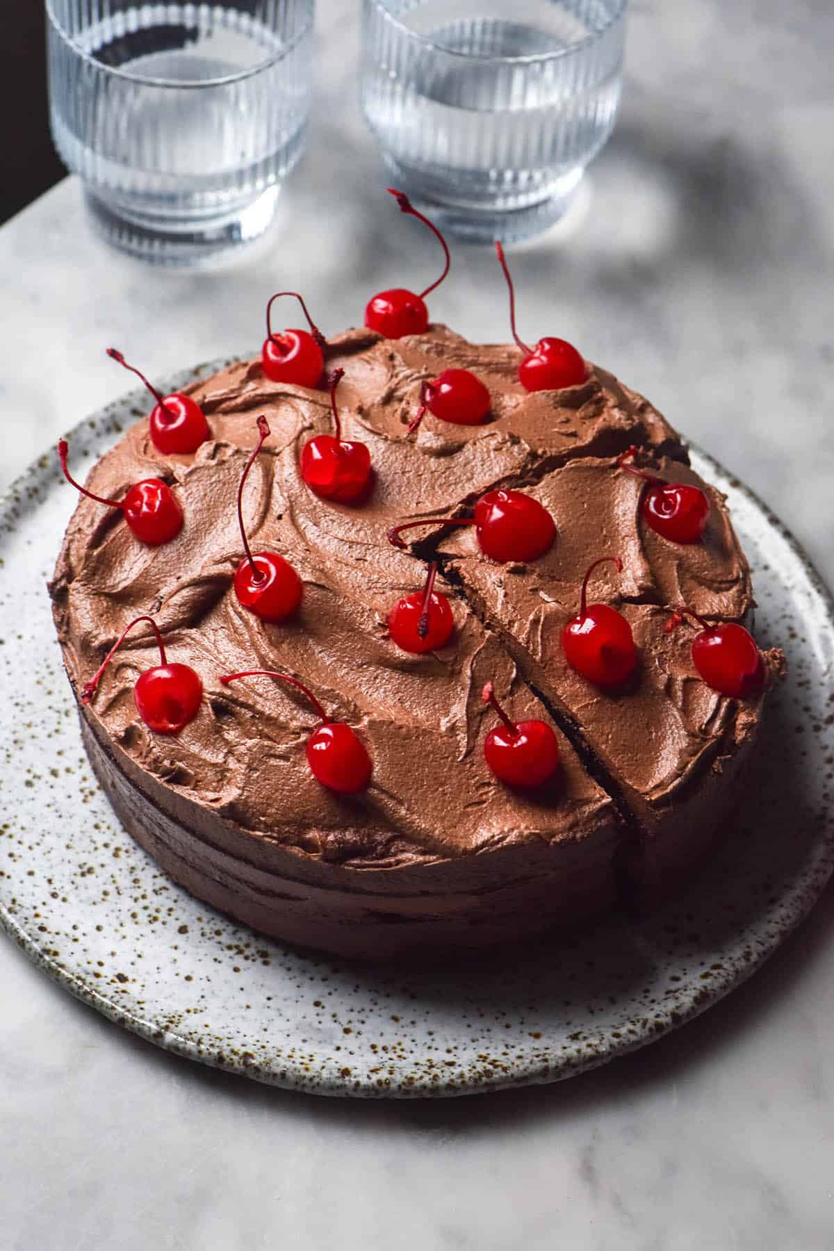 An aerial image of a gluten free dairy free chocolate layer cake topped with chocolate buttercream and maraschino cherries on a white marble table. Two glasses of water sit to the top right of the image. 