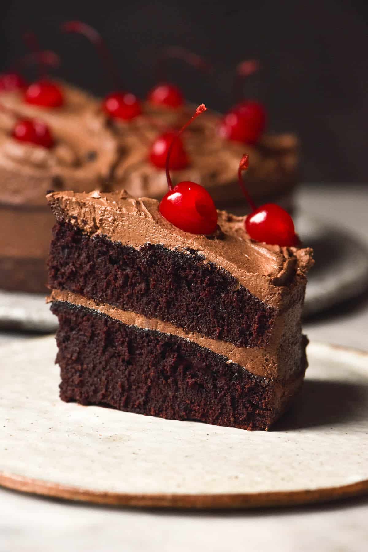 A side on image of a slice of gluten free dairy free chocolate layer cake topped with maraschino cherries. The slice sits atop a white plate on a white marble table. The whole cake sits behind the slice.