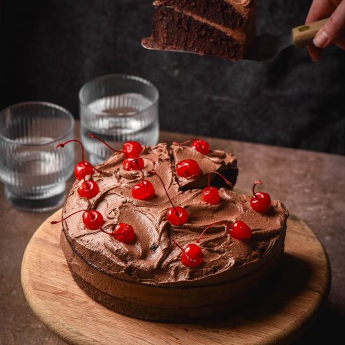 A side on image of a gluten free dairy free chocolate layer cake topped with dairy free chocolate buttercream and maraschino cherries. The cake sits atop a dark grey stone backdrop against a dark backdrop. A hand extends from the side of the image to take a slice of the cake.