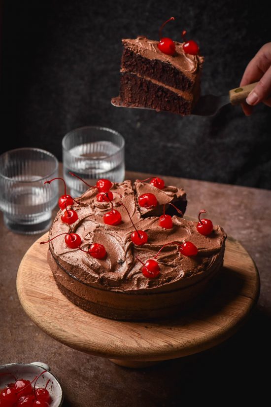 A side on image of a gluten free dairy free chocolate layer cake topped with dairy free chocolate buttercream and maraschino cherries. The cake sits atop a dark grey stone backdrop against a dark backdrop. A hand extends from the side of the image to take a slice of the cake.