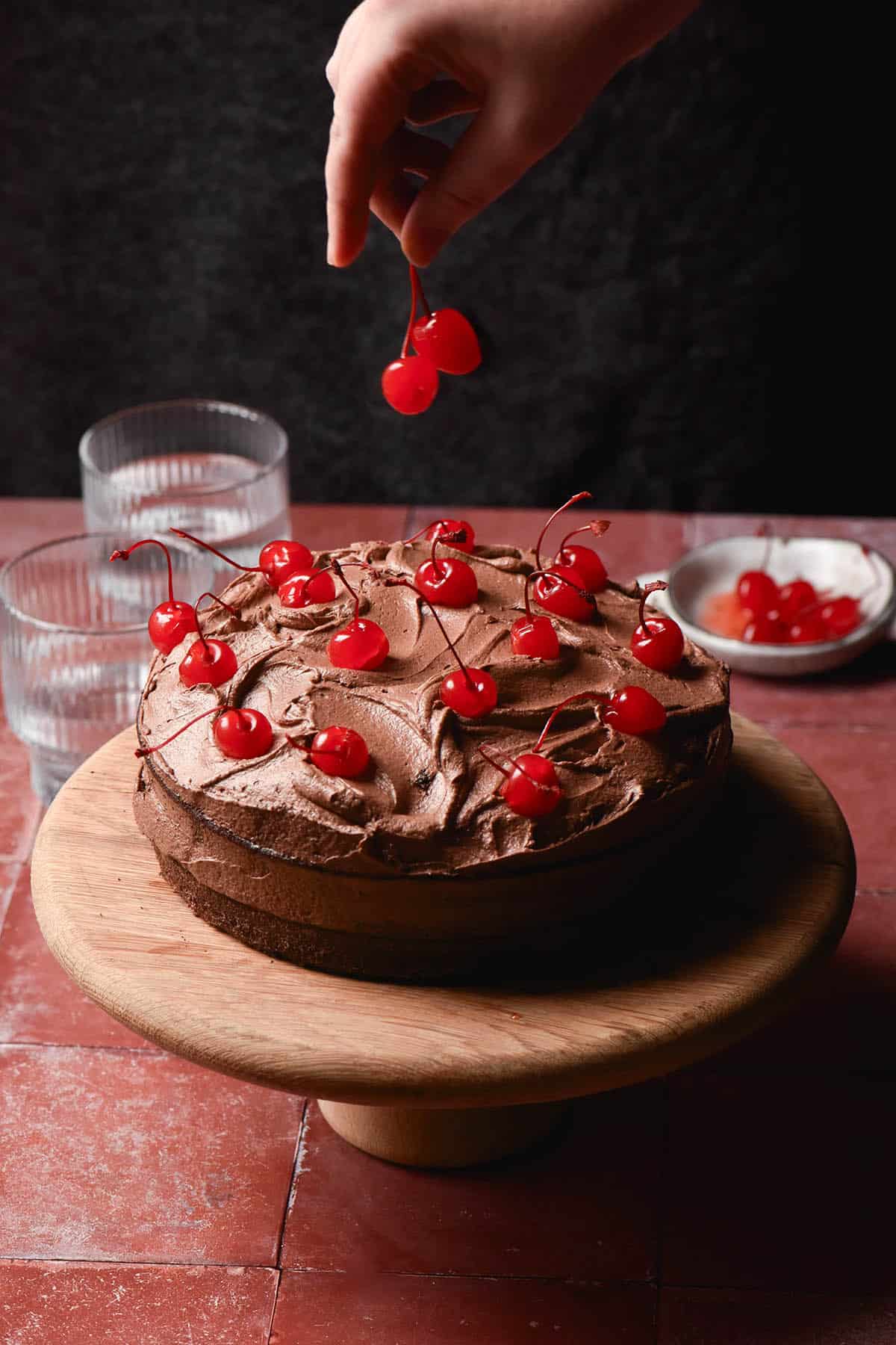 A side on image of a gluten free dairy free chocolate layer cake topped with dairy free chocolate buttercream and maraschino cherries. The cake sits atop a terracotta stone backdrop against a dark backdrop. A hand extends from the top of the image to add extra cherries onto the cake.