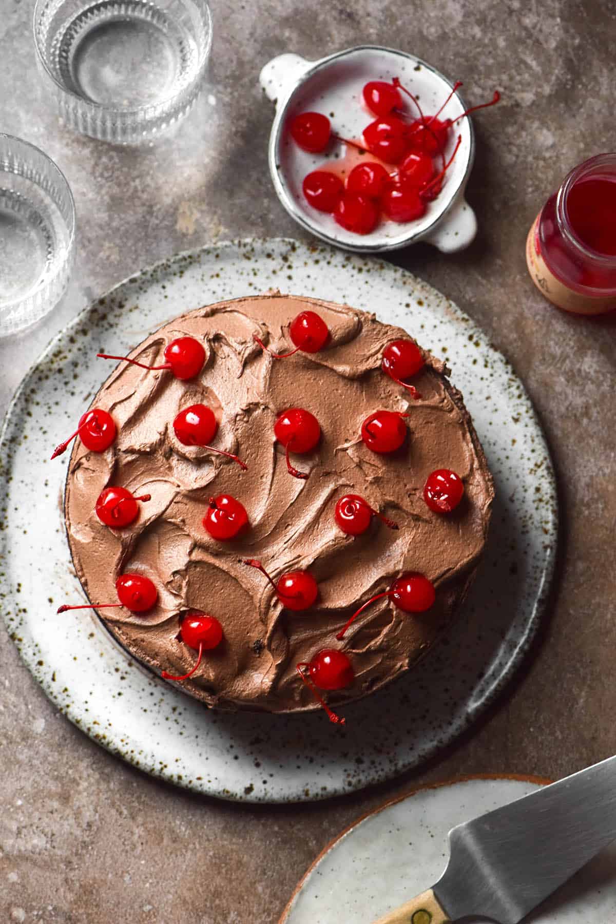 An aerial image of a gluten free dairy free chocolate layer cake topped with chocolate buttercream and maraschino cherries. The cake sits atop a stone backdrop and is surrounded by glasses of water, an extra plate and more maraschino cherries