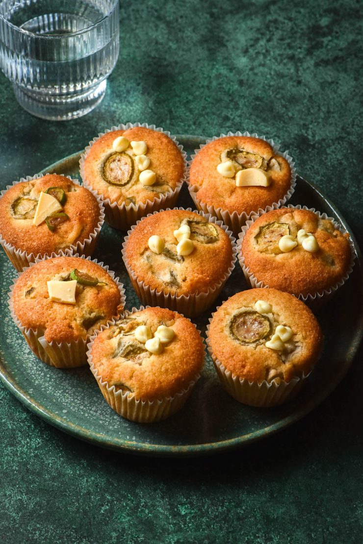 A side on image of a turquoise green plate topped with gluten free feijoa muffins which are golden brown and studded with white chocolate. The plate sits atop a turquoise green backdrop and a glass of water sits to the top left of the image.
