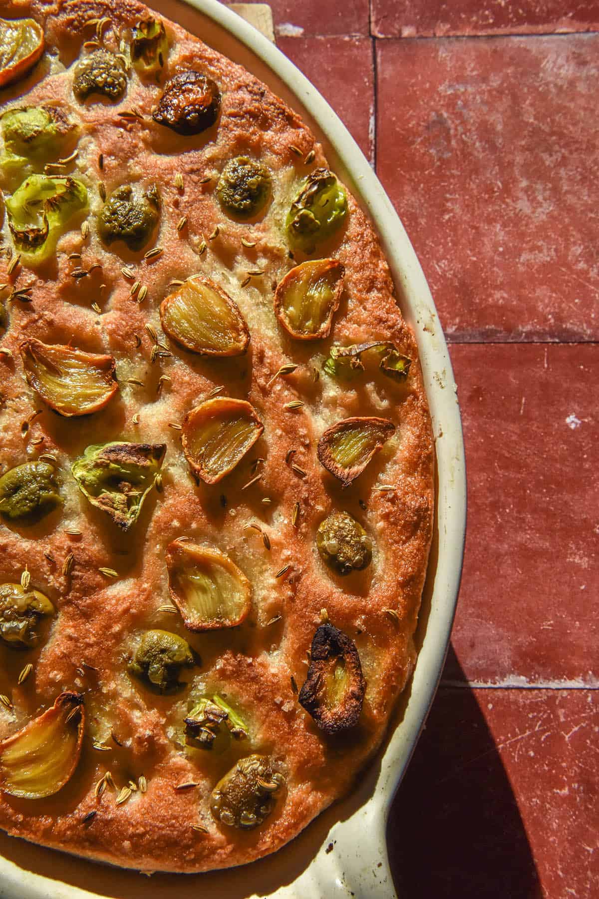 An aerial image of a golden brown gluten free focaccia topped with pickled roasted garlic, olives, pickled peppers and fennel seeds. The focaccia sits in a white ceramic baking dish atop terracotta tiles