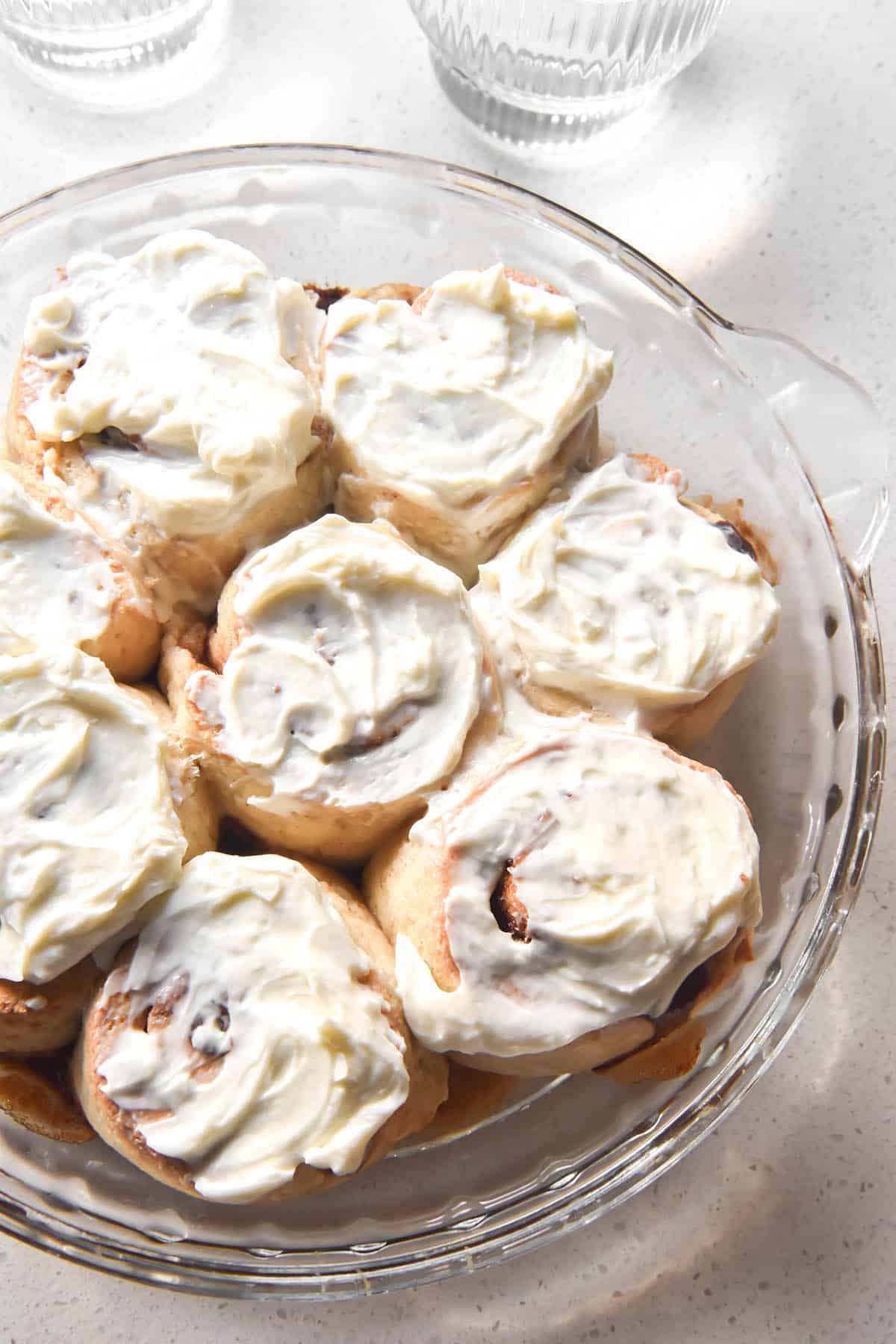 An aerial image of a pie plate filled with gluten free cinnamon rolls topped with cream cheese icing atop a white marble table.