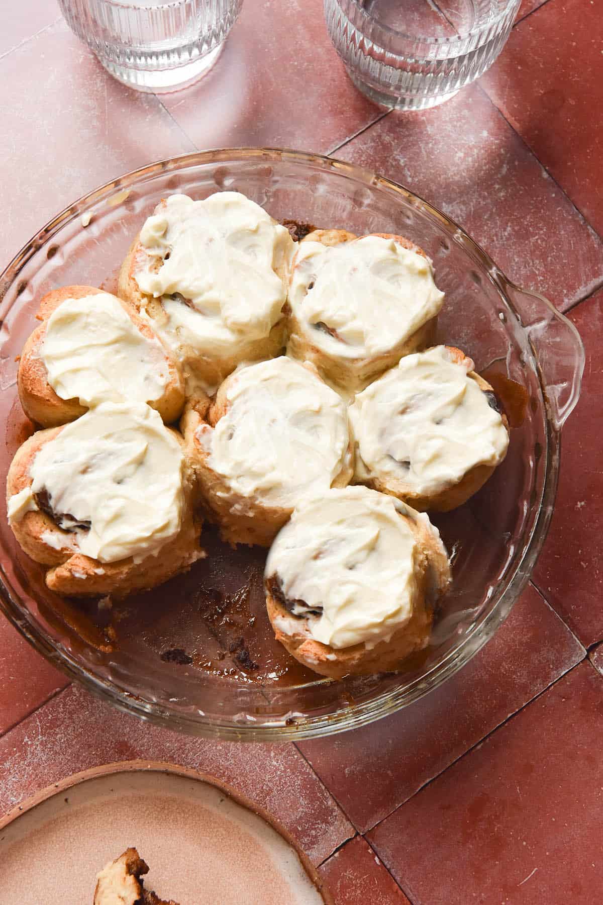 An aerial image of a plate plate filled with gluten free cinnamon rolls topped with cream cheese icing. The plate sits atop a terracotta tile backdrop and glasses of water sit to the top right of the pie plate.