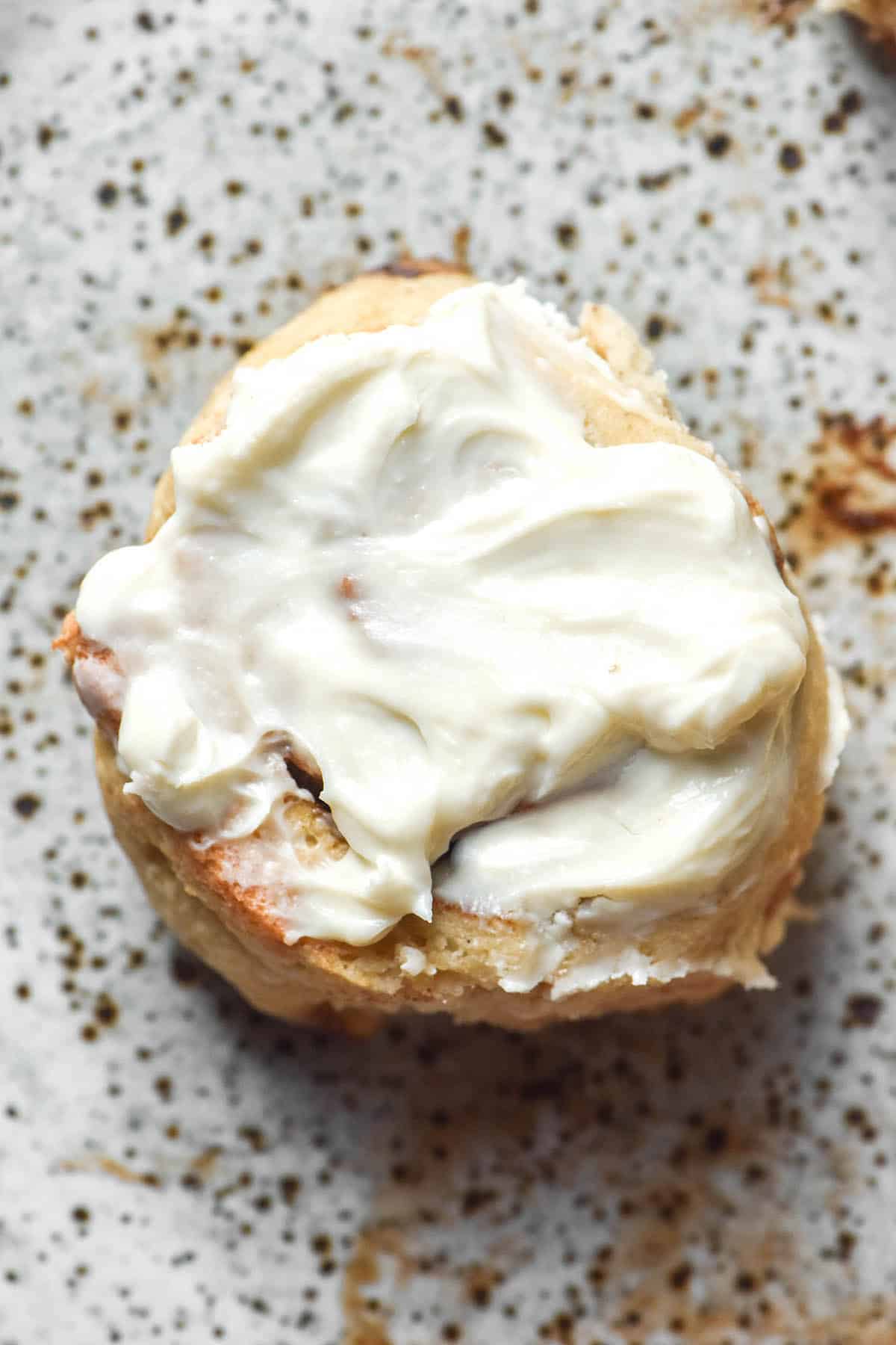 A close up aerial image of a gluten free yeast free cinnamon roll on a white speckled ceramic plate. The bun is golden brown and smothered in cream cheese icing.