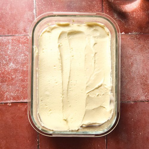 An aerial view of a glass container filled with lactose free cream cheese on a terracotta tile backdrop. Two glasses of water sit to the top right of the image