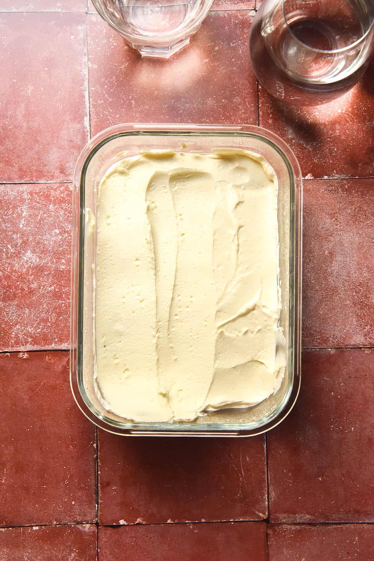 An aerial view of a glass container filled with lactose free cream cheese on a terracotta tile backdrop. Two glasses of water sit to the top right of the image