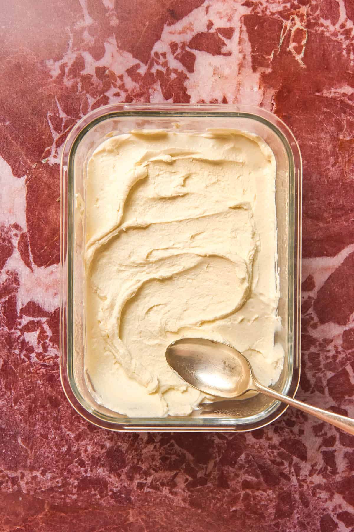 An aerial view of a glass container of lactose free cream cheese on a red marble backdrop. A spoon sits casually in the bottom right corner of the cream cheese container.