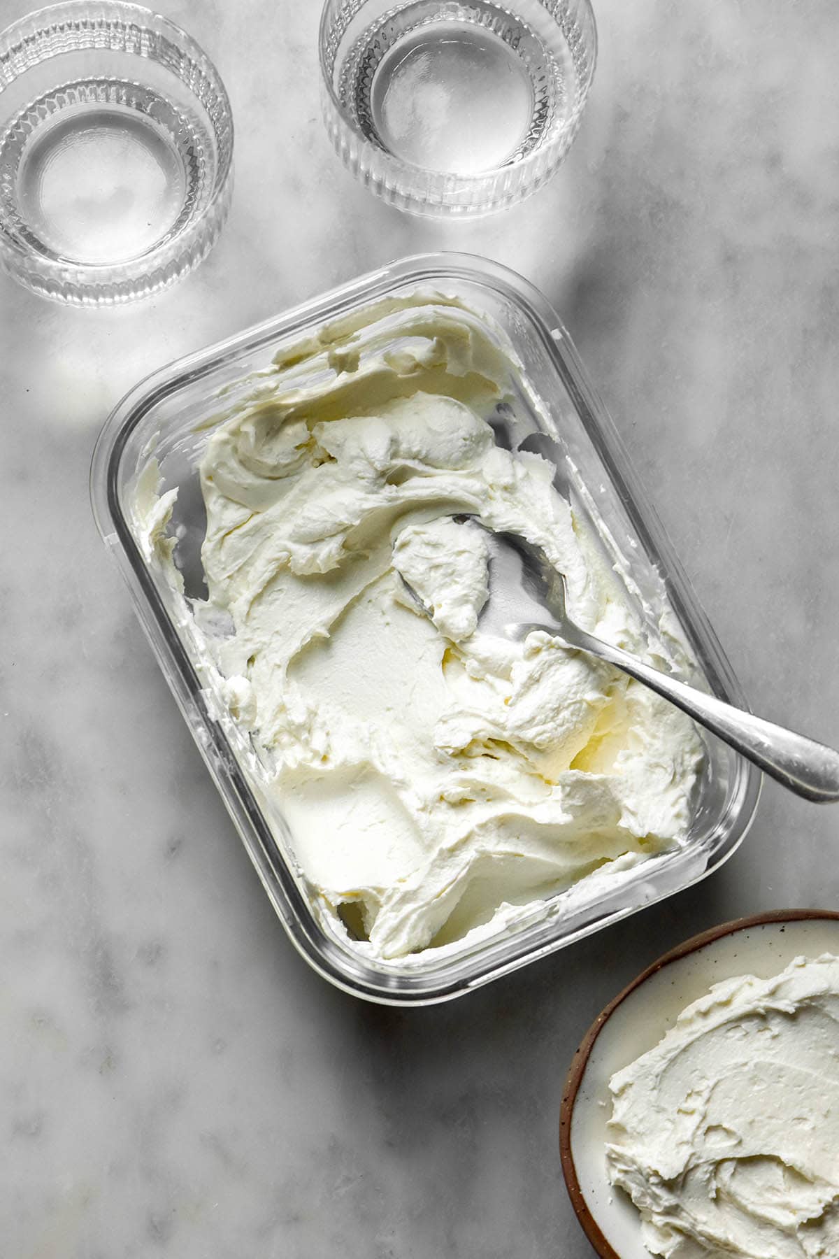 An aerial image of a glass container filled with lactose free cream cheese on a white marble table. A spoon sits casually in the container, while a small white bowl of cream cheese sits to the bottom right. Two sunlit glasses of water sit in the top left of the image