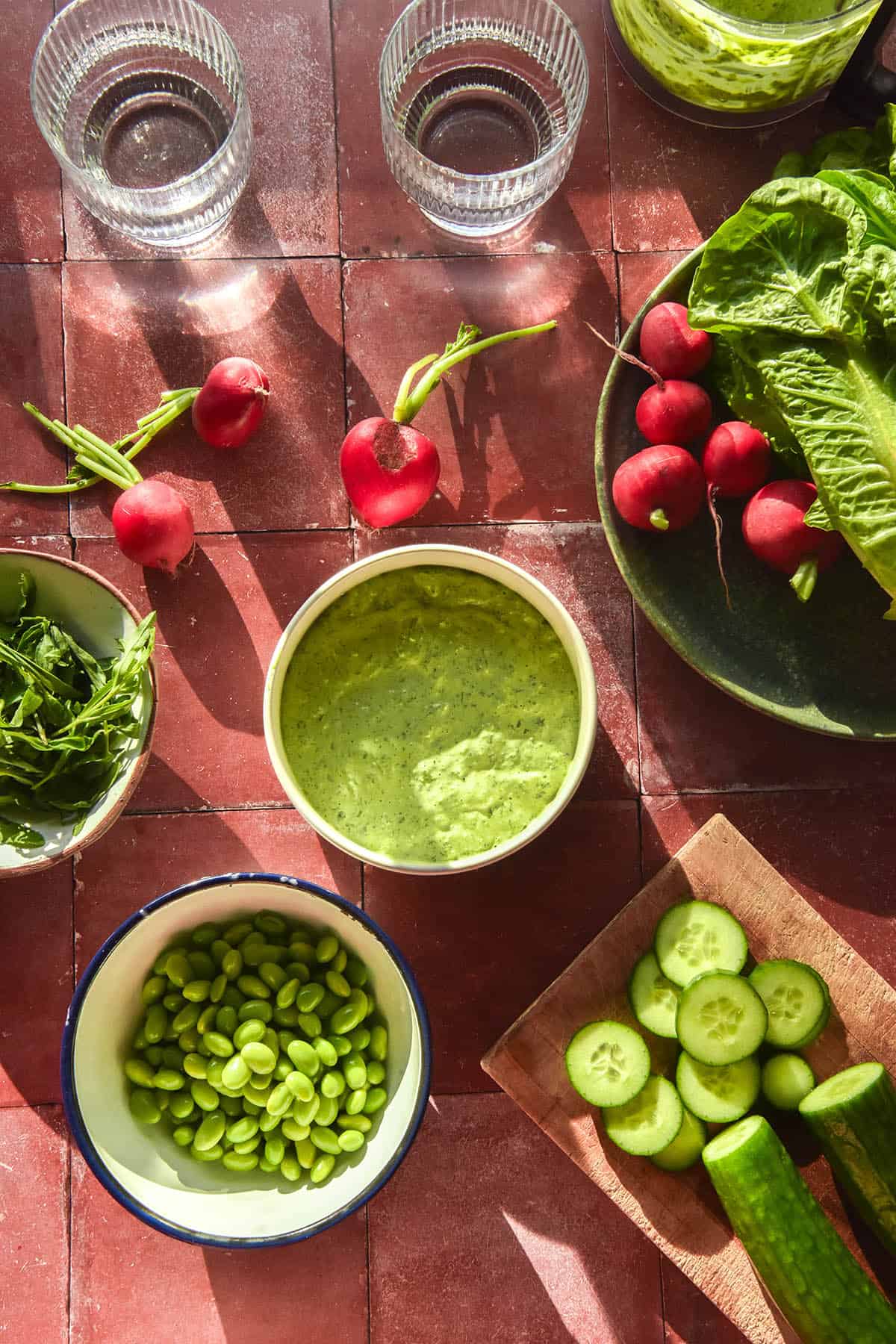 A sunlit aerial view of a bowl of low FODMAP Green Goddess salad dressing on a terracotta tile backdrop. The dressing is surrounded by ingredients for a salad and sunlit glasses of water.