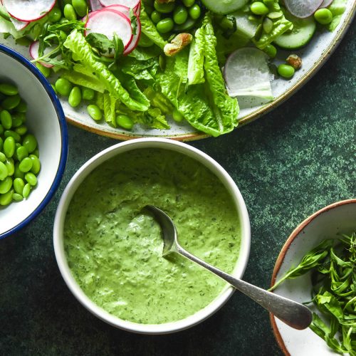 An aerial image of a bowl of Green Goddess dressing on a dark green backdrop. The dressing is surrounded by extra bowls of ingredients and a large salad topped with radishes and peas.