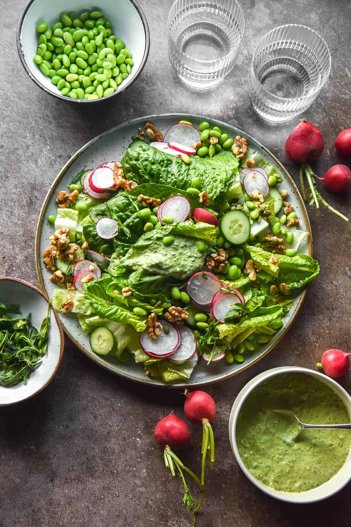An aerial image of a salad topped with low FODMAP Green Goddess dressing. The salad sits atop a medium grey backdrop and is surrounded by more dressing, water glasses and ingredients for the salad.