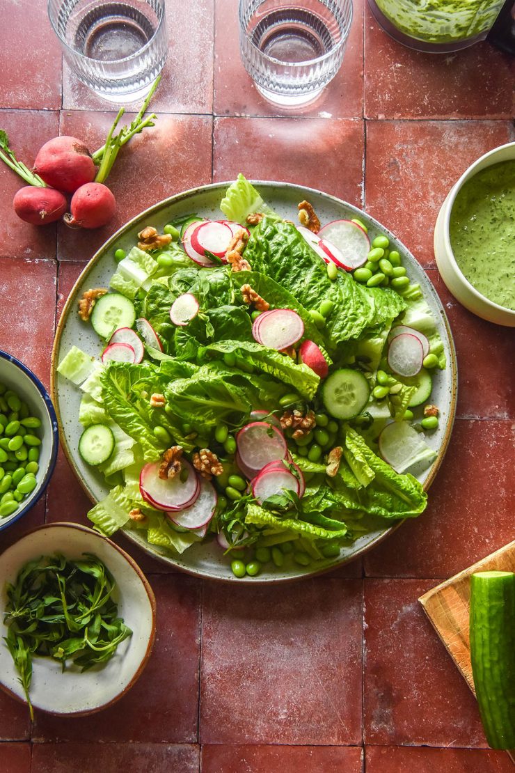 An aerial image of a salad ready to be topped with low FODMAP Green Goddess dressing. The salad sits atop a terracotta tile backdrop and is surrounded by a bowl of Green Goddess dip, glasses of water and ingredients for the salad.