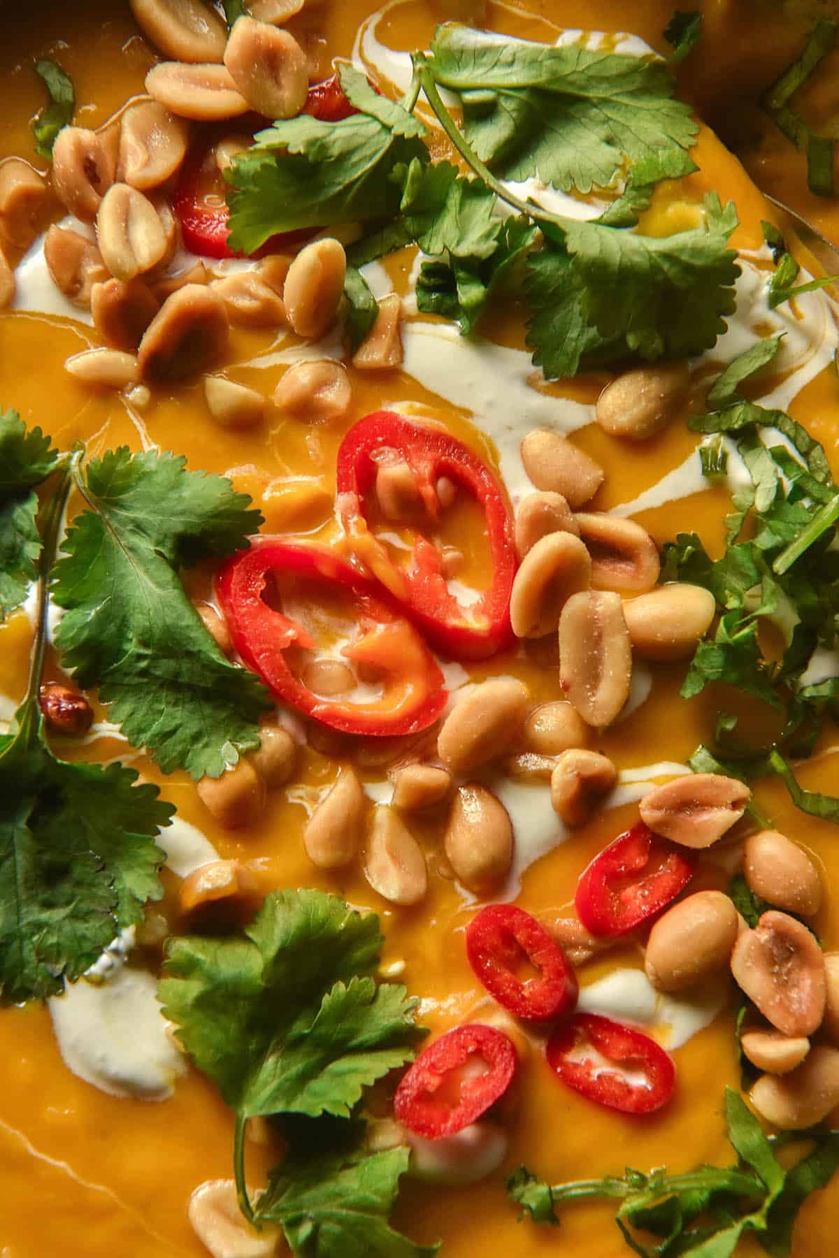 An aerial close up image of a bowl of Thai pumpkin soup topped with coconut cream, chopped chilli, coriander and toasted peanuts